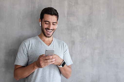Indoor daylight picture of handsome man wearing gray casual t-shirt, laughing happily being amused by content on screen of smartphone he is holding in both hands, standing against textured wall