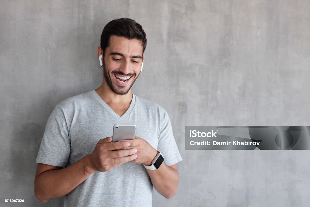 Indoor daylight picture of handsome man wearing gray casual t-shirt, laughing happily being amused by content on screen of smartphone he is holding in both hands, standing against textured wall Men Stock Photo