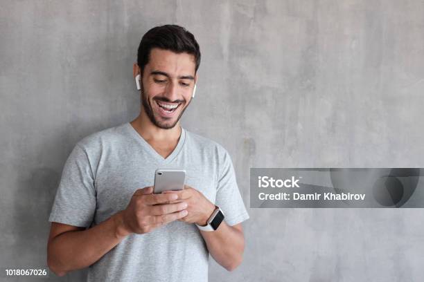 Indoor Daylight Picture Of Handsome Man Wearing Gray Casual Tshirt Laughing Happily Being Amused By Content On Screen Of Smartphone He Is Holding In Both Hands Standing Against Textured Wall Stock Photo - Download Image Now