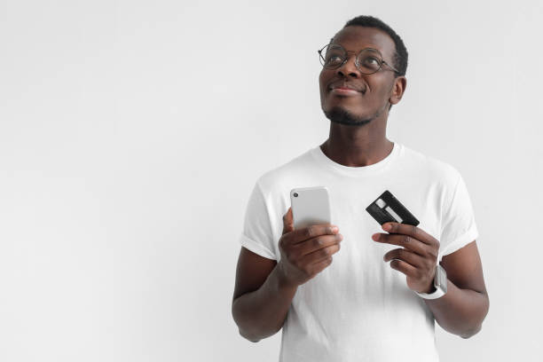 Closeup photo of young African male in eyeglasses pictured isolated on gray background thinking and dreaming about pleasant plans with credit card and smartphone in hands, ready to purchase in web Closeup photo of young African male in eyeglasses pictured isolated on gray background thinking and dreaming about pleasant plans with credit card and smartphone in hands, ready to purchase in web ticket photos stock pictures, royalty-free photos & images