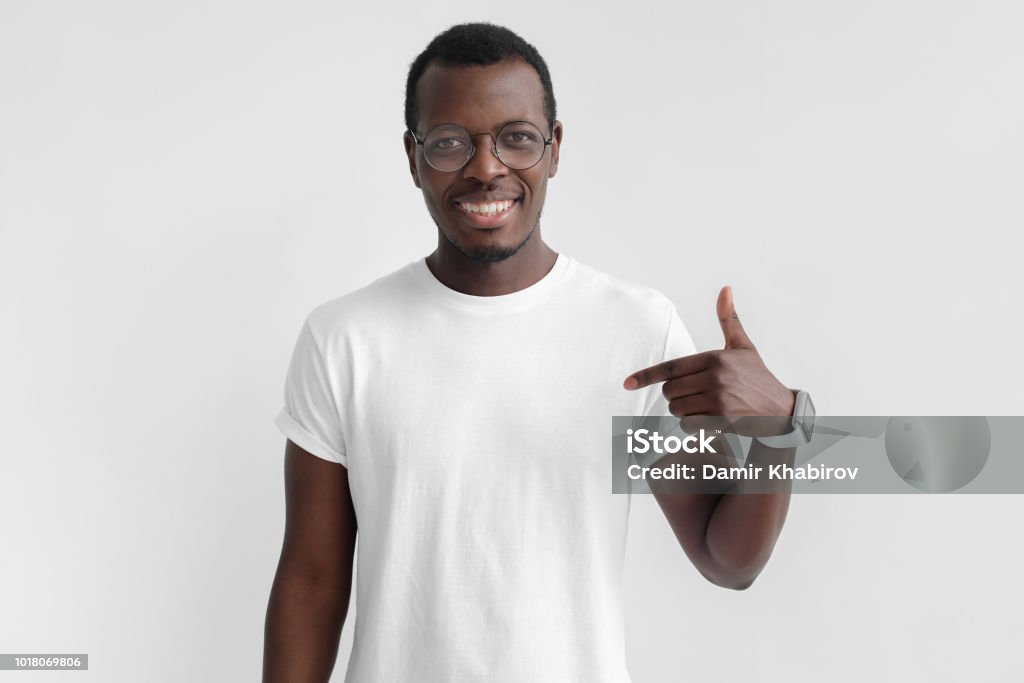 Indoor photo of young African American man pictured isolated on grey background pointing to his white blank T-shirt drawing attention to advertisement on it, promoting goods, apps or services T-Shirt Stock Photo