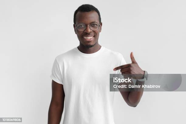 Foto de Foto Interior Da Jovem Afroamericano Homem Retratado Isolado No Fundo Cinza Apontando Para Sua Camiseta Branca Em Branco Chamando A Atenção Para O Anúncio Nele Promovendo Produtos Aplicativos Ou Serviços e mais fotos de stock de Camiseta