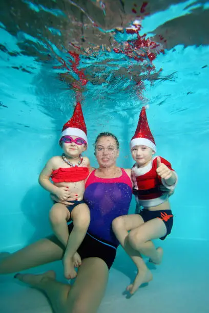 Photo of Family, mother, daughter and son in red cap of Santa Claus posing underwater at the bottom of the pool, looking at me and smiling. Portrait. Shooting under water. Vertical orientation