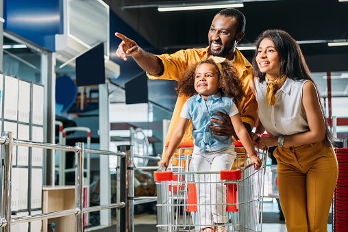 smiling african american man pointing by finger to little daughter and wife in supermarket