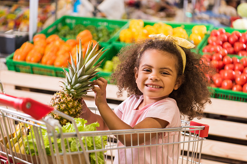 smiling african american kid sitting in shopping trolley with pineapple in grocery store