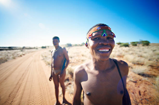 Portrait hunters Bushmen San Namibia standing on a sandy dirt road Mariental, Kalahari Desert, Namibia - May 26, 2018: Close-up portrait hunters Bushmen or San people standing next to a sandy road with the sun from behind. The San people, also known as Bushmen are members of various indigenous hunter-gatherer peoples of Southern Africa bushmen stock pictures, royalty-free photos & images