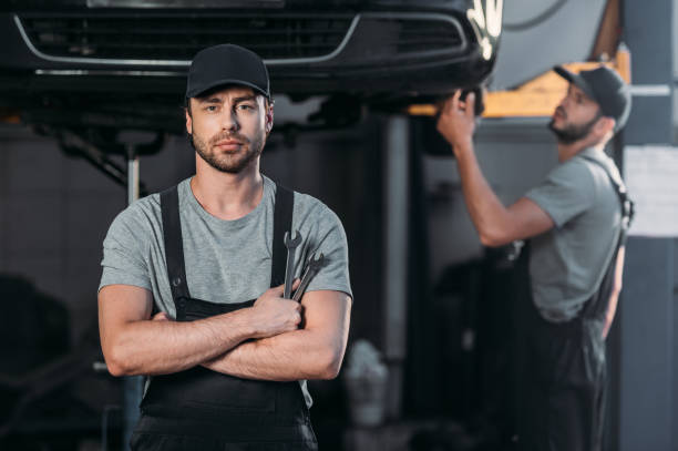 istock auto mechanic posing in overalls with crossed arms, while coworker working in workshop behind 1018158554