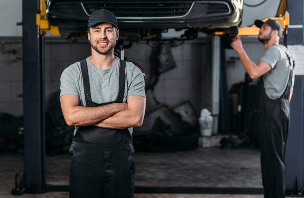 istock smiling mechanic in overalls posing with crossed arms, while colleague working in workshop behind 1018158560