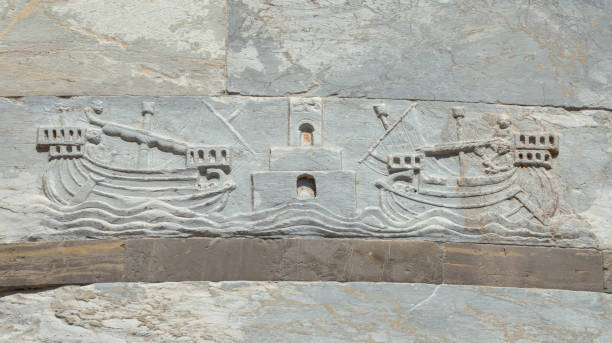 Pisa, Italy - May 11, 2018: Navigation and maritime trade in the Middle Ages. Two ships with sailors enter the Port of Pisa Old Maritime Republic with lighthouse. Medieval relief on Leanign Tower wall (12th century)
