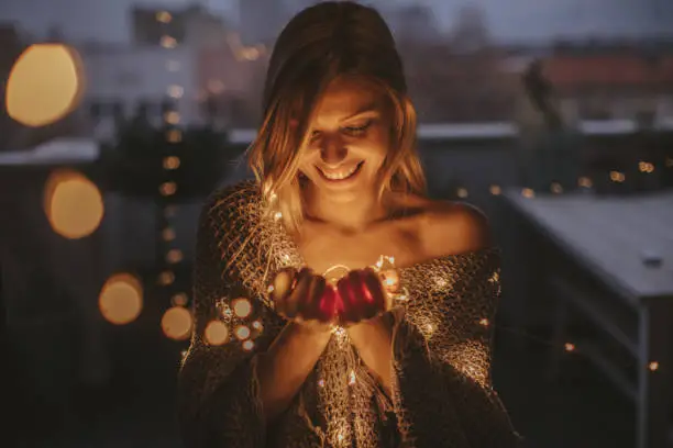 Young, smiling woman standing outdoors on the decorated terrace of her apartment and holding string lights
