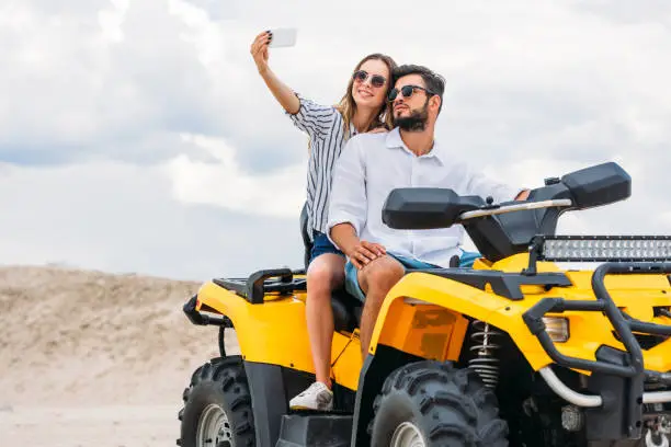 attractive young couple taking selfie while sitting on ATV in desert