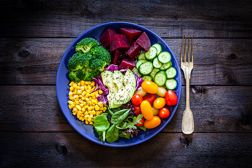 Top view of a blue salad plate filled with fresh organic colorful vegetables shot on rustic dark wooden table. Vegetables included in the composition are tomatoes, cucumber, beet, broccoli, corn, avocado, arugula and lettuce. This ingredients are typical of the Mediterranean cuisine. Low key DSRL studio photo taken with Canon EOS 5D Mk II and Canon EF 100mm f/2.8L Macro IS USM.