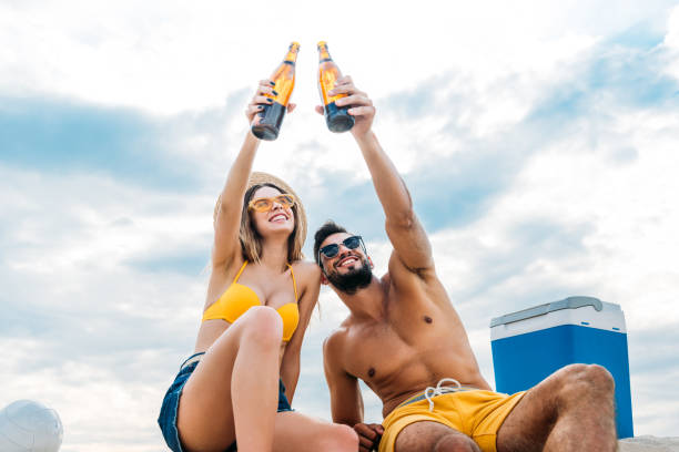 нижний вид красивой молодой пары поднимая бутылки пива перед облачным небом, сидя на песке - shirtless beach women bikini стоковые фото и изображения