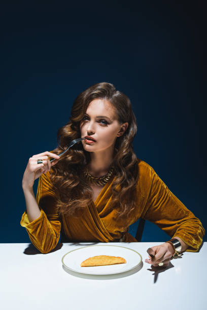 portrait of woman in luxury clothing with fork sitting at table with cheburek on plate with blue backdrop - blue plate fotos imagens e fotografias de stock