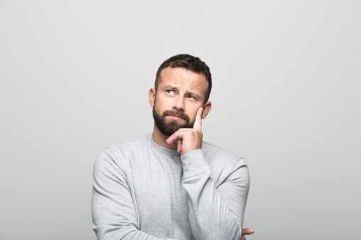 Portrait of pensive bearded young man looking away with hand on chin. Studio shot, grey background.