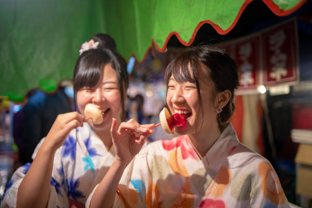 Young female friends eating 'Anzu Ame' - Japanese apricot candy at 'Yatai' night market Young female friends eating 'Anzu Ame' - Japanese apricot candy at 'Yatai' night market yukata photos stock pictures, royalty-free photos & images