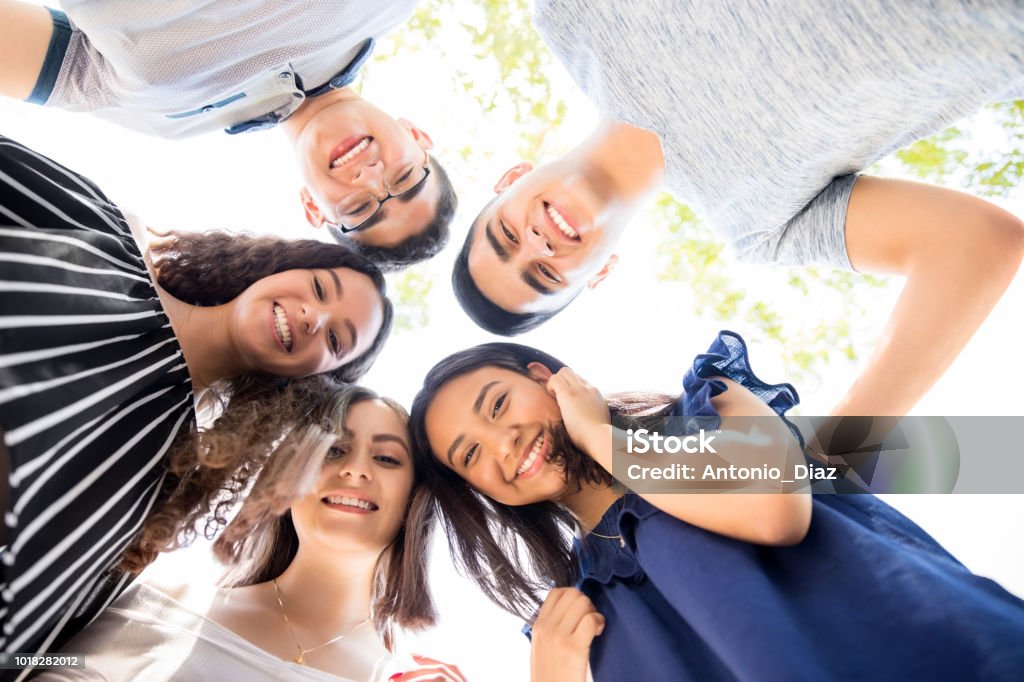 Friends standing in a circle outdoors Low angle view of group of teenagers standing in a huddle with their heads touching and looking at camera Teenager Stock Photo