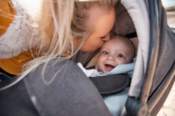 A close up shot of a mother kissing her son on the head, the baby boy is in his stroller and the woman is crouching down next to him.