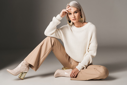 attractive fashionable woman posing in white trendy sweater, beige pants and autumn heels, on grey