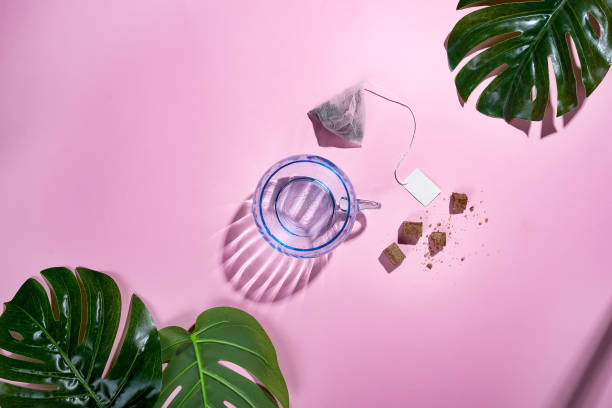 Top view, office table desk with copy space. Workspace frame with green tropical leaf and tea cup, tea bag and sugar on pastel pink background with beautiful deep shadows. Flay lay.