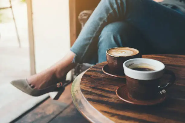 Closeup image of two cups of hot latte coffee on vintage wooden table with woman sitting in background
