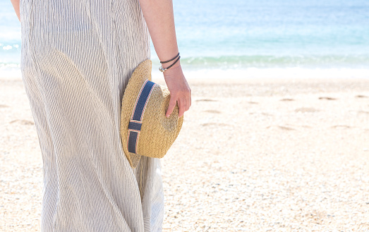 A mid section of a rear view girl in light dress holding a straw sun hat and on a sandy beach with copy space