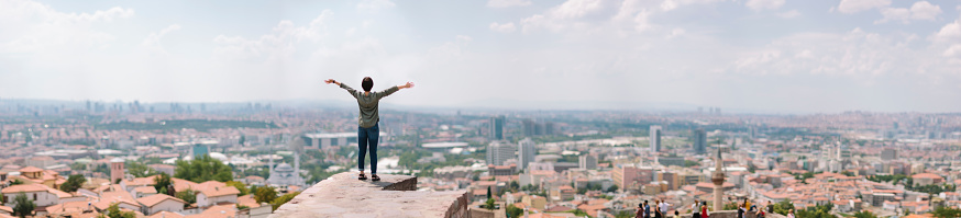 A young Japanese female traveller is looking at the panoramic scene of Ankara with her arms raised in the air