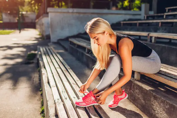 Young woman athlete tying sneakers sitting on bench on sportsground in summer. Preparation for training. Workout