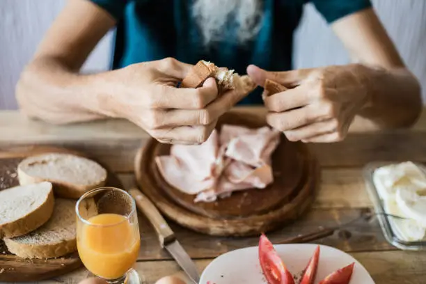 Man tearing bread slice, having breakfast. Close-up, selective focus. Fruit juice, tomato, mortadella and bread slices in background
