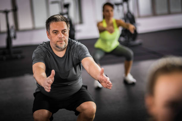 overweight middle-aged man doing squats in aerobics class - women sweat healthy lifestyle exercising imagens e fotografias de stock