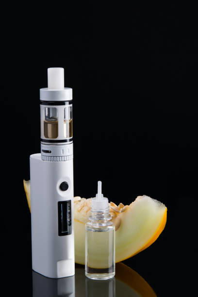 istock white e-cigarette with melon flavor dressing, on the background of ripe yellow fruit, with reflection on the black background. 1018666972