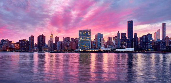 A panoramic view of Midtown Manhattan, New York from Gantry Plaza State Park. Reflection of city lights and the color spectrum of the blue hour on the East River. Pink and blue cotton candy clouds are flowing above the office buildings. Pink sparkles reflect the lights on the river surface.