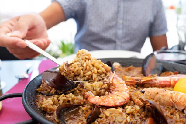 closeup of a young caucasian serving a typical spanish seafood paella from the paellera, the paella pan, placed on a table set for lunch