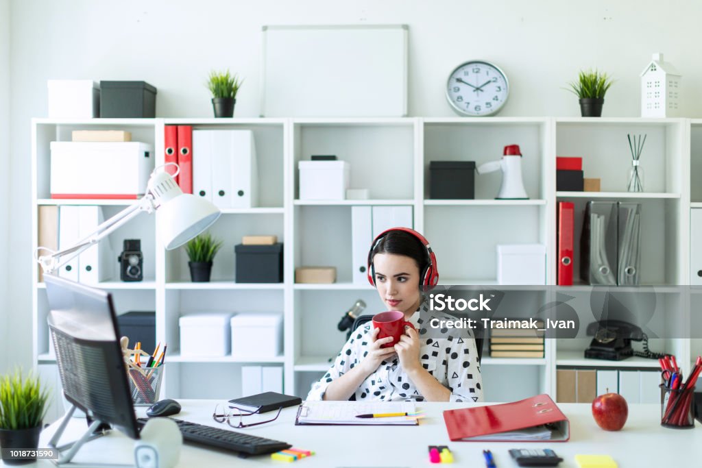 A young girl sits in headphones at a table in the office, holds a red cup in her hands and looks at the monitor. Beautiful young girl in a white blouse in a geometric pattern and blue jeans. Girl with dark hair and beautiful make-up. A charming young girl is working in a bright office. photo with depth of field. Making Stock Photo