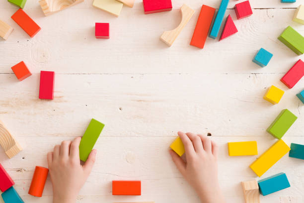 top-ansicht auf kinderhände spielen mit bunten holzziegeln auf weißem tisch hintergrund. junge gebäude mit holzkonstrukteur. bildungskonzept - cube baby child block stock-fotos und bilder