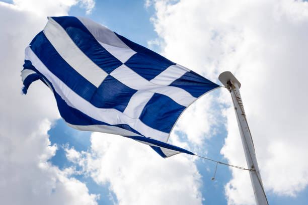 istock waving Greek flag on a flagpole against a blue sky with clouds 1018747204