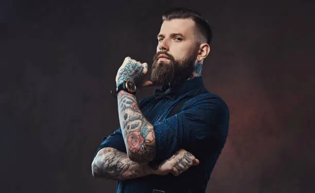 Pensive handsome old-fashioned hipster in a blue shirt and suspenders, standing with hand on chin in a studio. Isolated on a dark background.