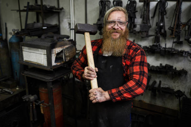 istock Positive confident strong male blacksmith in protective eyewear and apron holding big hammer for shaping metal and looking at camera in workshop 1018801204