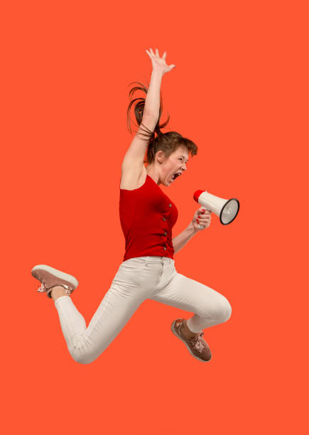 istock Beautiful young woman jumping with megaphone isolated over red background 1018838532