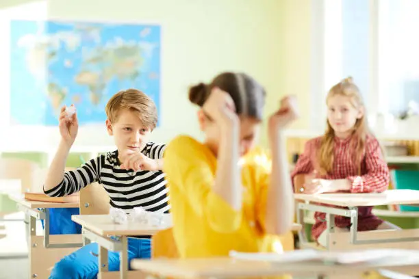 Angry school bully throwing paper ball at new girl and sitting at desk in geography classroom, bullying at school concept