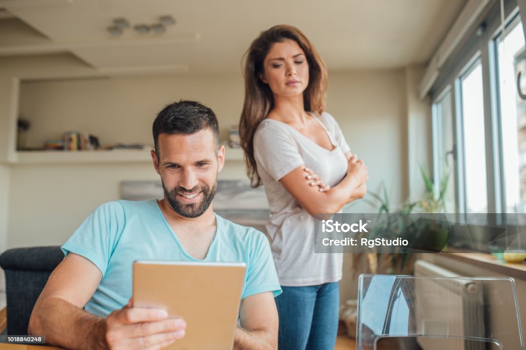 Young woman jealously looking at the smiling man using digital tablet Couple having relationship issues Envy Stock Photo