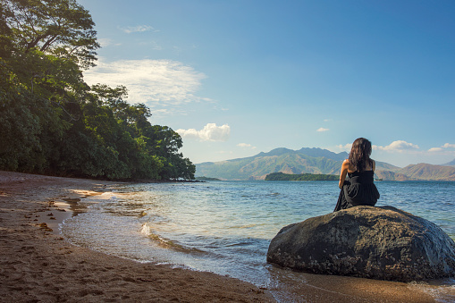 A woman sits on a rock looking at the view by a quiet beach on the west coast of Luzon, Philippines.