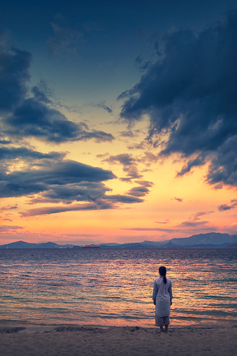 A woman stands alone on a beach looking out towards horizon in the evening.