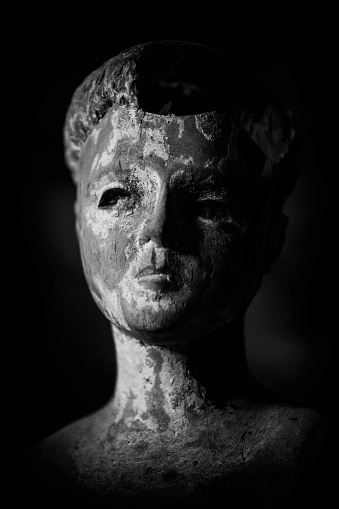 Black and white image of a weathered and damaged wooden figure, with a missing piece in its head.