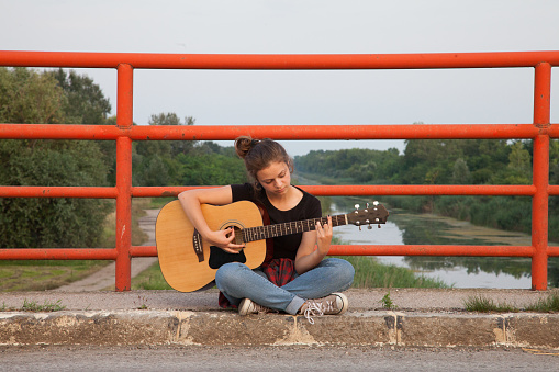 Teenage girl is sitting on a bridge and playing an acoustic guitar.
