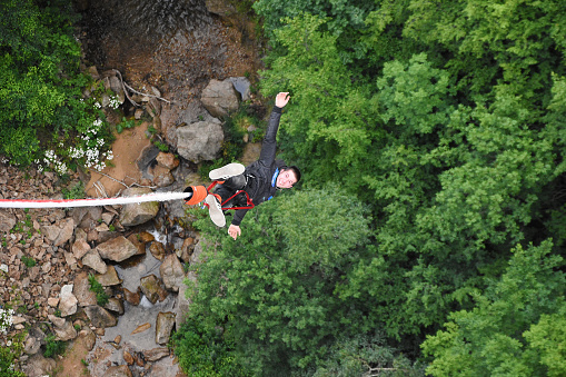 Klisura Bridge, Bulgaria - June 23, 2018: Young smiling boy hanging on a rope during bungee jump from 70 meters high bridge. Out of focus mountain river, rocks and green trees seen at background