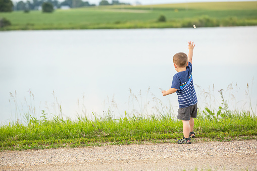 Rear view of a two year old boy throwing a rock into the water from up above on a rural gravel driveway. The rock can be seen in mid-air, as it has just left the boy's hand.