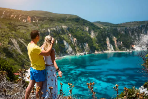 Photo of Birthday gift. Young man closing hes girlfriend eyes in front of gorgeous seascape panorama of blue lagoon. Couple love. Turquoise Petani beach on Kefalonia island Greece