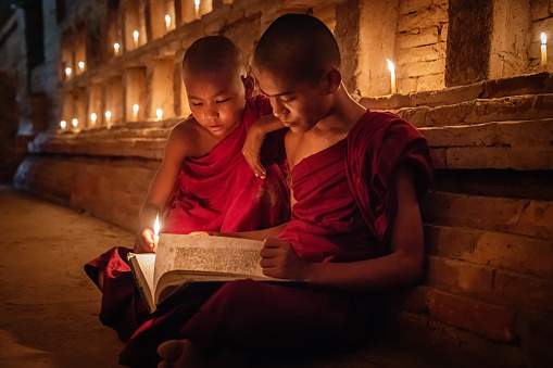 Two young burmese buddhist novice monks in their typical religious clothing sitting side by side inside candle lit temple tomb together studying a buddhist book. Natural Candle Light Peaceful Scene. Bagan, Mandalay Region, Myanmar.