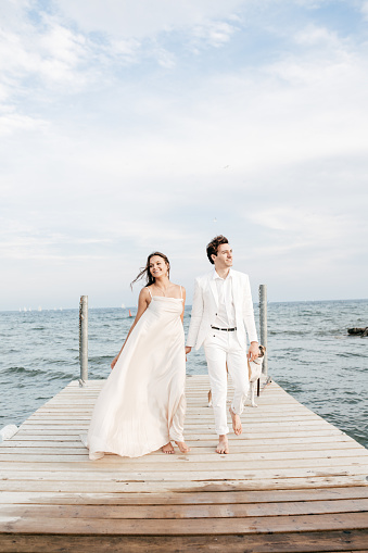Wedding portrait. A brunette bride in a long dress and a groom in a classic suit are standing, embracing, in love against the background of a lake.
